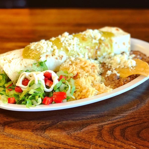 A burrito covered in green sauce and cheese, served with rice, refried beans, and a side salad.