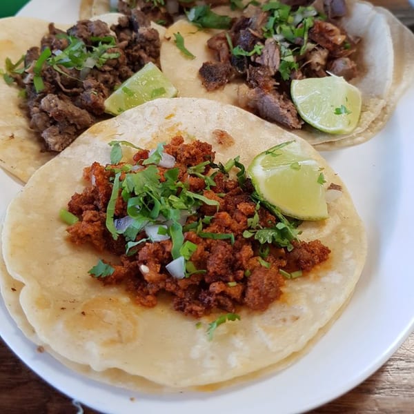 Three Mexican street tacos with various meat fillings, cilantro, onions, and lime wedges on a white plate.