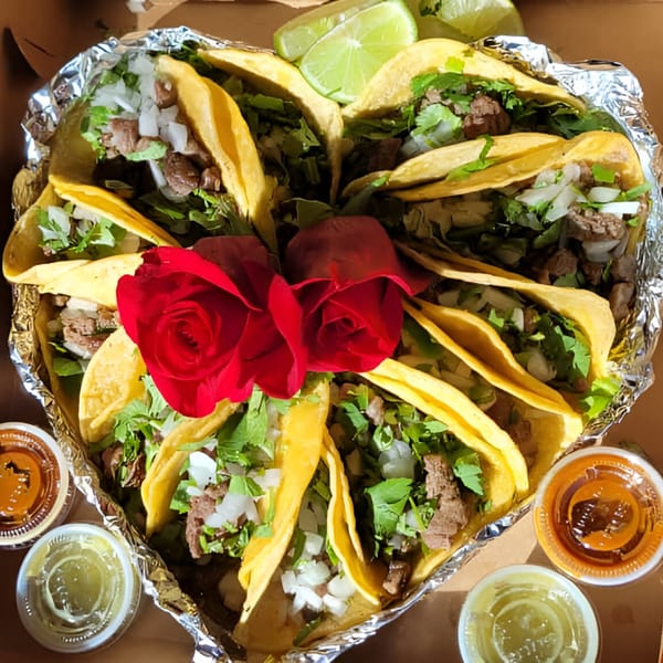 Heart-shaped arrangement of tacos with meat, onions, cilantro, lime wedges, and two red roses.