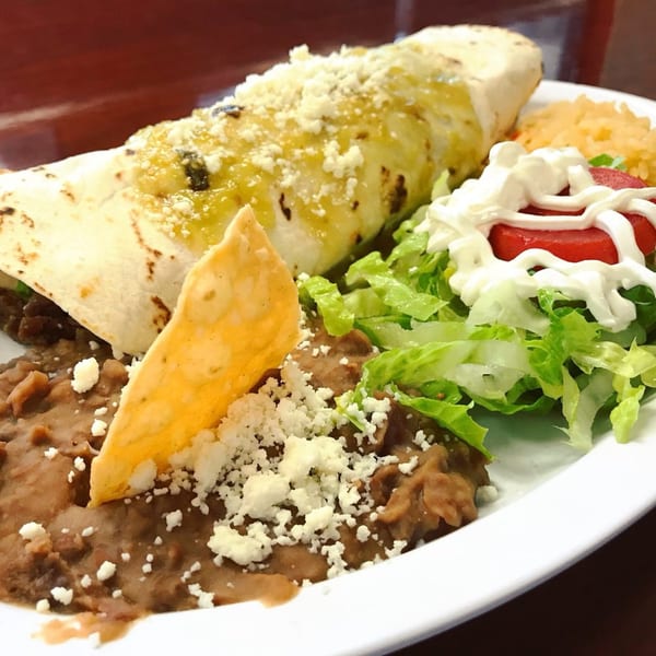 A large burrito topped with green sauce and cheese, served with refried beans, rice, and salad.